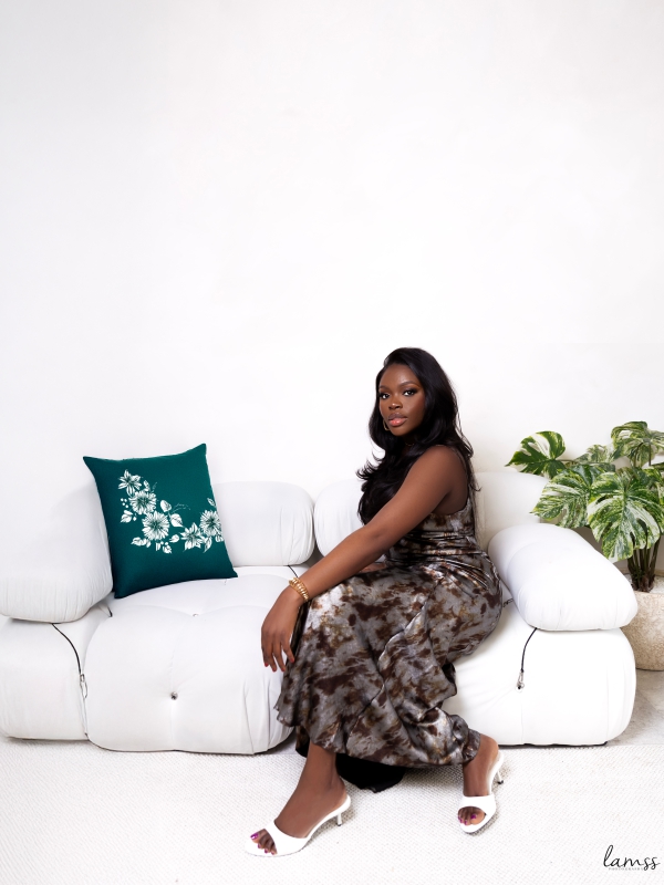 A woman in a patterned brown dress sitting on a white sofa with a green decorative pillow and a plant nearby. — portrait photography London by Lamss Media